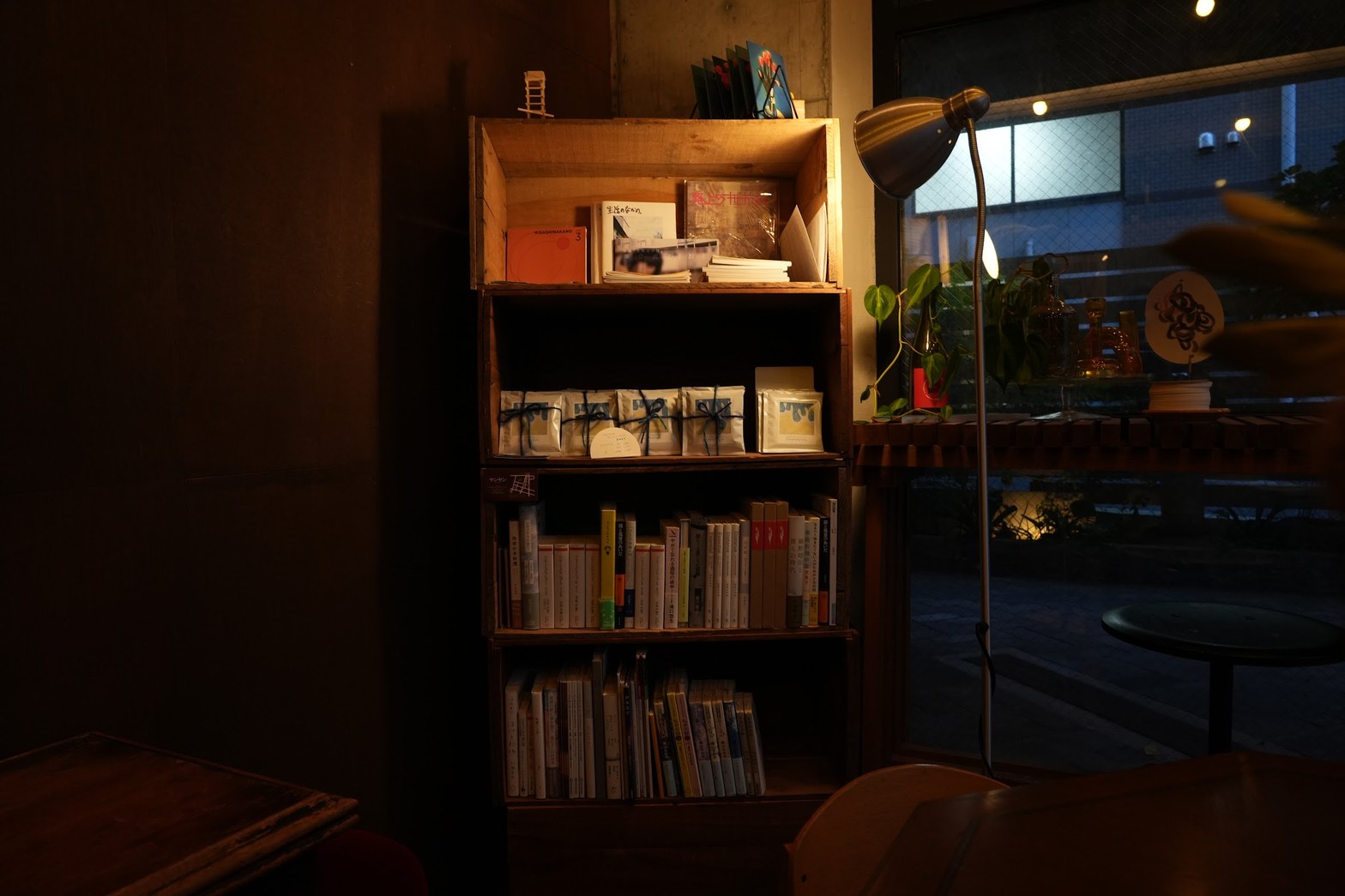 木製棚の中段にリボン掛けのコーヒー豆パッケージが並び、上段に雑誌や冊子、下段に文庫本が詰まった店内の本棚。右手に真鍮色のフロアランプが灯り、窓越しに夜の外光が差し込む。