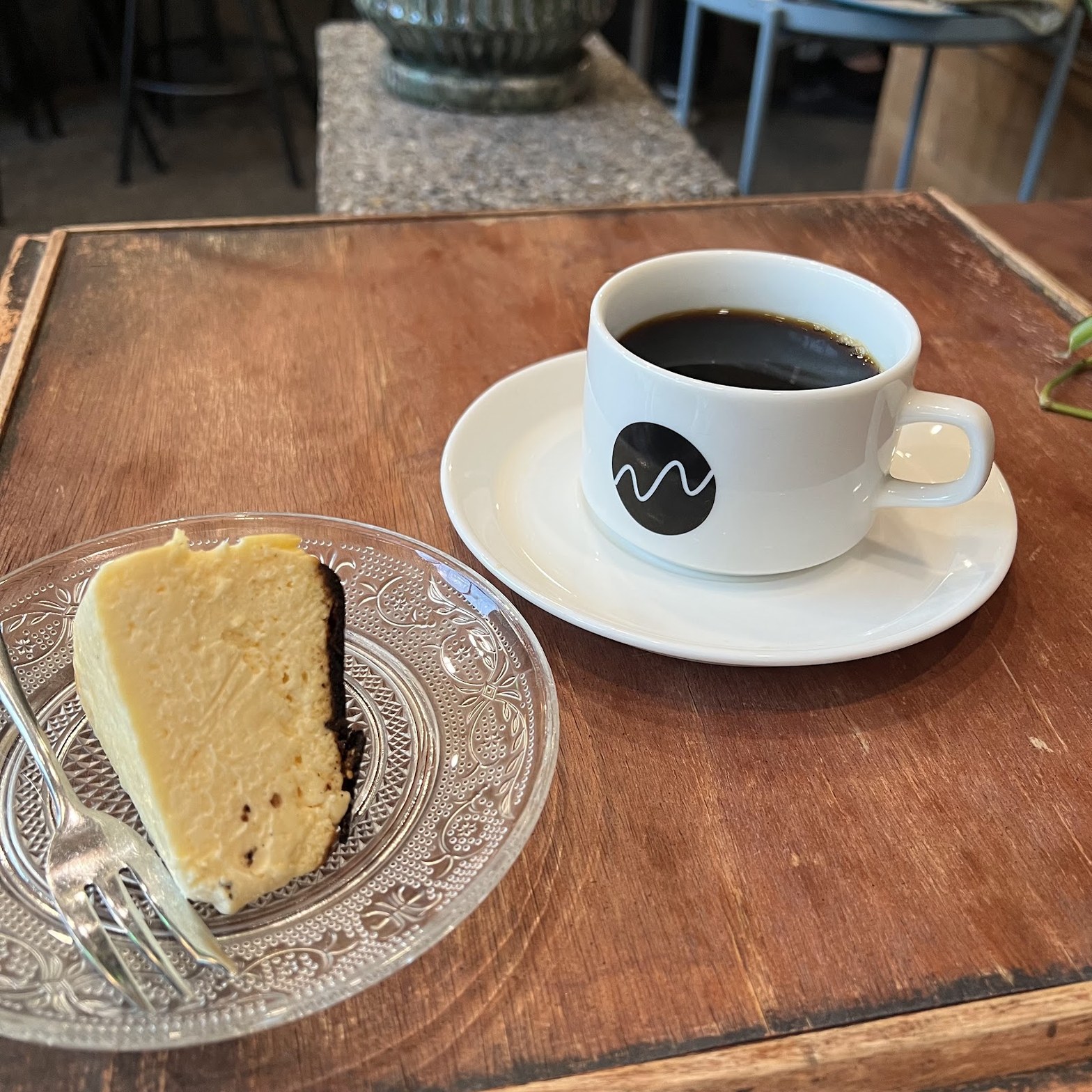 白いカップにロゴ入りのブレンドコーヒーと、ガラス皿に乗ったバスクチーズケーキのひと切れ。木目のテーブルに並んで置かれている。